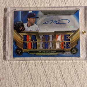 Peut inclure: Carte de baseball avec Jacob deGrom, lanceur. La carte a un design bleu et or avec les mots "DAYTIME DOMINANCE" en grandes lettres. Elle est enfermée dans un étui de protection transparent. La carte est marquée "1/3" et comprend un autographe.