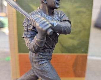 May include: A bronze-colored baseball player figurine in a batting stance, holding a bat. The figure wears a cap and a pinstriped uniform. The base of the figurine has the number 4. The background includes a vintage-style card.