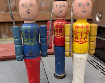 Op de afbeelding: Drie houten speelgoed soldaten met geschilderde gezichten en hoeden. De soldaat links draagt een blauw en rood uniform, de middelste soldaat draagt een rood en blauw uniform en de soldaat rechts draagt een geel en wit uniform.