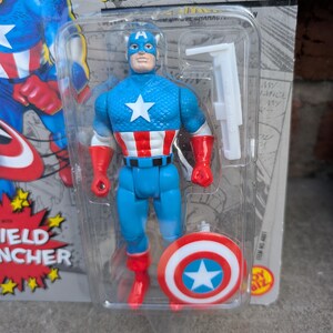 Puede incluir: Figura de acci&oacute;n del Capit&aacute;n Am&eacute;rica en un paquete de pl&aacute;stico. La figura es azul con una estrella blanca en el pecho, guantes y botas rojas, y un escudo a juego. El embalaje dice "SUPER HEROES" y "CAPTAIN AMERICA".