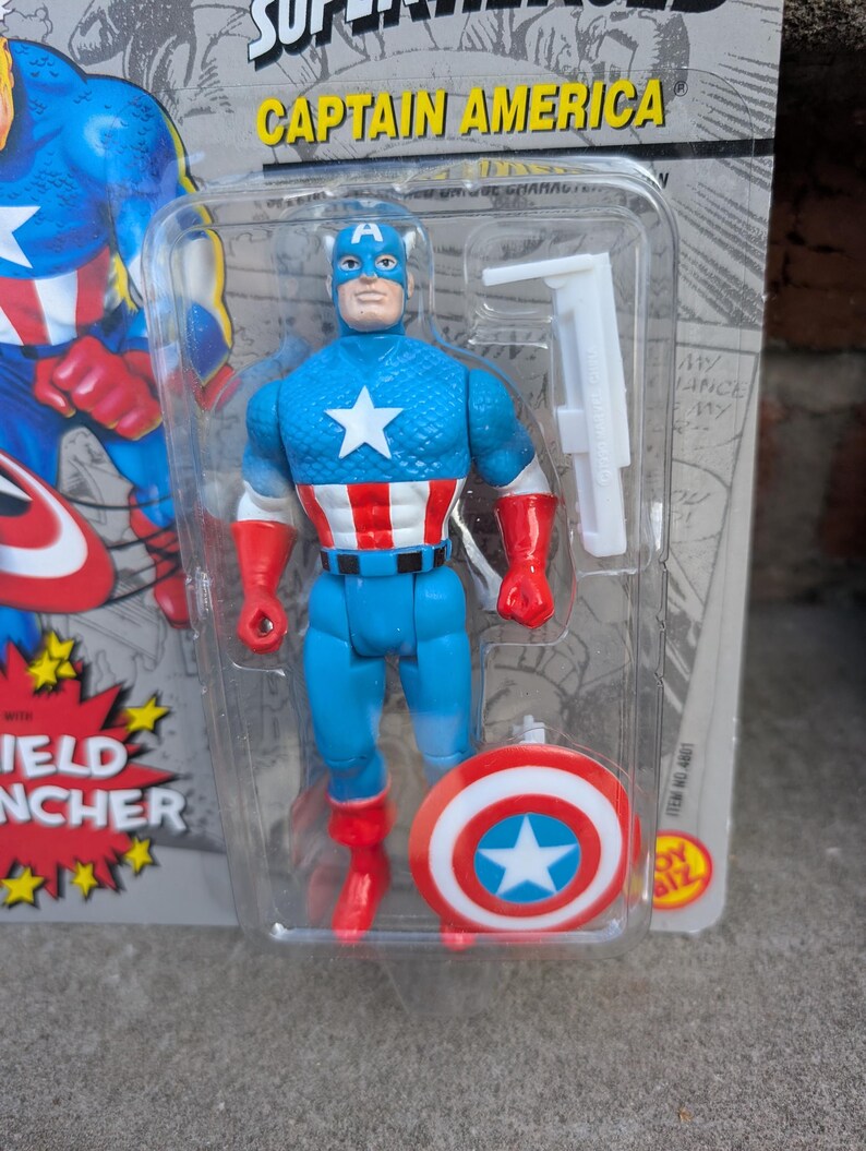 Puede incluir: Figura de acci&oacute;n del Capit&aacute;n Am&eacute;rica en un paquete de pl&aacute;stico. La figura es azul con una estrella blanca en el pecho, guantes y botas rojas, y un escudo a juego. El embalaje dice "SUPER HEROES" y "CAPTAIN AMERICA".