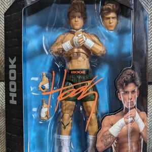 Könnte beinhalten: Eine Wrestling-Actionfigur von Hook aus der Unmatched Collection von AEW. Die Figur trägt ein grünes und schwarzes Wrestling-Outfit mit einem schwarzen und orangefarbenen Gürtel, auf dem "HOOK" steht. Die Figur ist in einer Kampfhaltung mit erhobenen Händen dargestellt. Die Figur befindet sich in einer durchsichtigen Plastikvitrine mit blauem Hintergrund. Die Figur ist Teil der Serie 7 und für Kinder ab 8 Jahren geeignet.