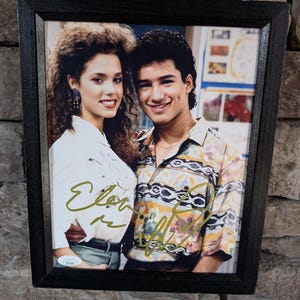 May include: Framed photograph featuring two smiling people, a woman and a man, posing together. The man wears a patterned shirt, and the woman wears a white blouse. Both have signatures in gold ink. The frame is black.
