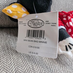 Puede incluir: Un peluche mini bean bag de Minnie Mouse. El peluche tiene una cabeza negra con un lazo amarillo de lunares, un vestido rojo de lunares y guantes blancos. La etiqueta dice "Mini Bean Bag Minnie".