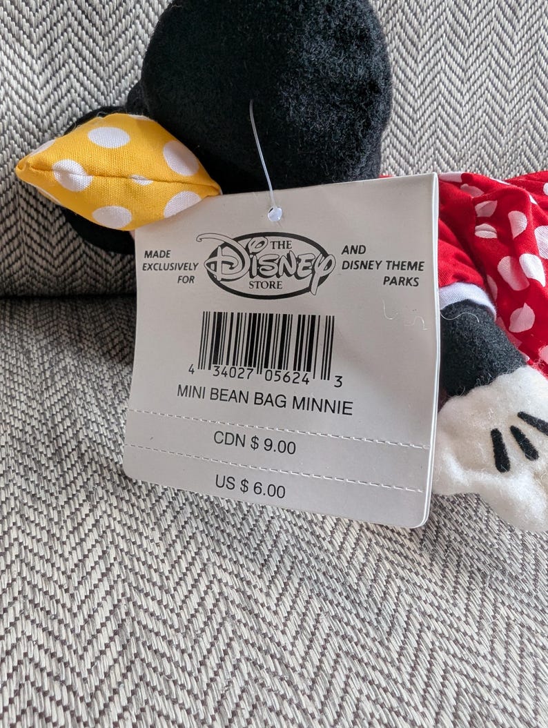 Puede incluir: Un peluche mini bean bag de Minnie Mouse. El peluche tiene una cabeza negra con un lazo amarillo de lunares, un vestido rojo de lunares y guantes blancos. La etiqueta dice "Mini Bean Bag Minnie".