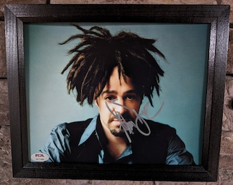 Puede incluir: Retrato enmarcado de un hombre con rastas, con una camisa azul y un chaleco negro. El retrato está firmado y tiene una pegatina de autenticación PSA. El marco es marrón oscuro y el fondo es azul claro.
