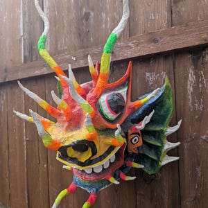 May include: A colorful, handcrafted dragon mask with a vibrant palette of red, orange, yellow, green, and blue. The mask features large white teeth, expressive eyes, and long, curved horns. It is mounted on a wooden fence.