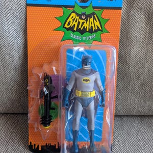 May include: Batman Classic TV Series action figure in packaging. The figure is gray and blue with a yellow utility belt and a bat symbol on the chest. The packaging is orange and blue with the Batman logo.