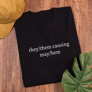 May include: Black t-shirt with white text that reads "they/them causing may/hem".