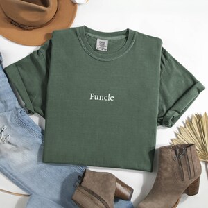 May include: An olive green t-shirt with the word "Funcle" printed in white. The shirt is laid flat with rolled-up sleeves, surrounded by blue jeans, brown boots, and a brown hat.