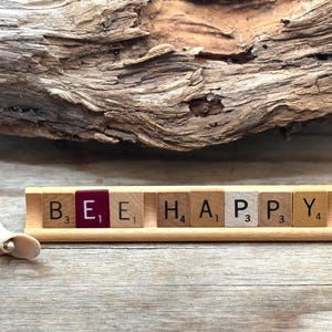May include: Wooden tile display spelling "BEE HAPPY" with individual letter tiles. The letter "E" is on a maroon tile. A small, off-white seashell sits to the left of the display. The background includes a piece of driftwood and a wooden surface.