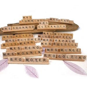 May include: Wooden Scrabble tiles arranged on wooden tile holders. The tiles spell out various names and words, including "Mrs." and surnames. The arrangement is on a white surface with decorative leaf accents, ideal for personalized gifts or home decor.
