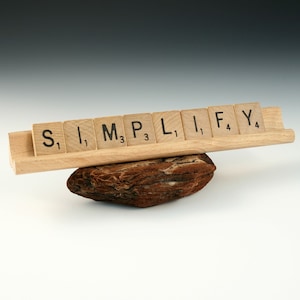 Puede incluir: Un soporte de madera para fichas de Scrabble con la palabra "SIMPLIFY" escrita. El soporte descansa sobre un trozo de madera marrón texturizada. Las fichas son de color marrón claro con letras y números negros.