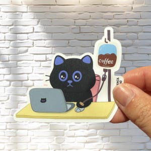 May include: A black cat with blue eyes sits at a desk with a laptop and a coffee IV bag labeled "coffee".