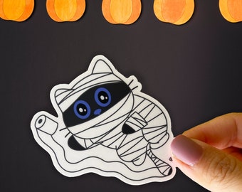 May include: A white and black cartoon cat dressed as a mummy with blue eyes. The cat is holding a roll of white bandage in its paw. The cat is on a white background.