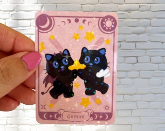 May include: A pink and purple holographic sticker featuring two black cats holding a gold star. The cats are wearing angel wings and devil horns. The sticker reads "Gemini" at the bottom.