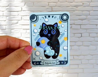 May include: A blue and white sticker featuring a black cat holding a blue pitcher with a yellow star. The cat has a green wreath around its head and blue eyes. The sticker reads "Aquarius" at the bottom.