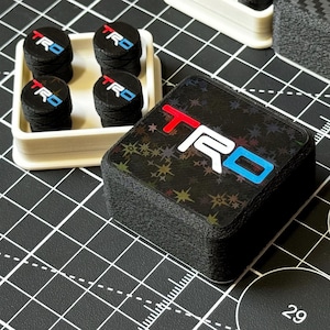 May include: A collection of black TRD logo accessories. The accessories include a stack of square-shaped items with the TRD logo in red, white, and blue, and a set of four round items with the same logo, displayed in a white tray.