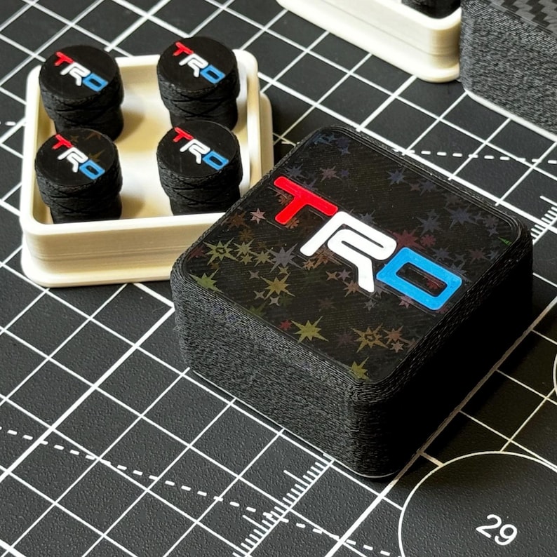 May include: A collection of black TRD logo accessories. The accessories include a stack of square-shaped items with the TRD logo in red, white, and blue, and a set of four round items with the same logo, displayed in a white tray.