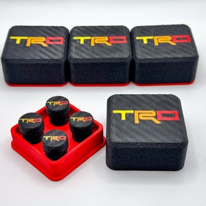 May include: Black and red TRD logo tire valve stem caps and matching square boxes. The caps and boxes have a carbon fiber texture. The TRD logo is yellow and red. The caps are in a red holder.