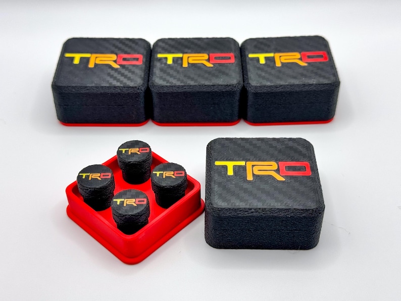 May include: Black and red TRD logo tire valve stem caps and matching square boxes. The caps and boxes have a carbon fiber texture. The TRD logo is yellow and red. The caps are in a red holder.