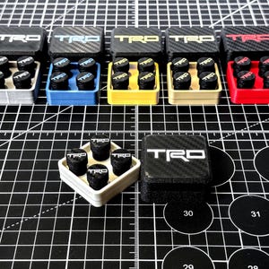 May include: A collection of TRD-branded tire valve stem caps in various colors, including silver, blue, yellow, beige, and red. Each set includes six black caps with white TRD lettering, presented in matching colored containers.