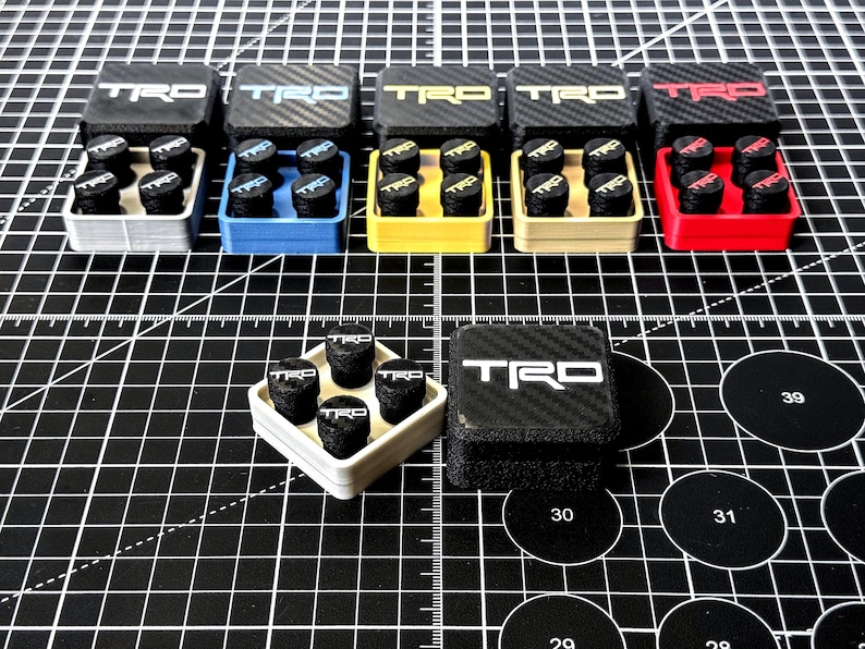 May include: A collection of TRD-branded tire valve stem caps in various colors, including silver, blue, yellow, beige, and red. Each set includes six black caps with white TRD lettering, presented in matching colored containers.