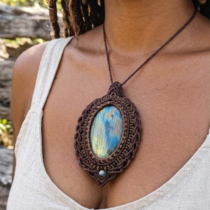 May include: A large oval pendant featuring a central iridescent blue and gold labradorite gemstone encased in intricate dark brown macrame cord work. The pendant hangs from a thin brown cord and includes a small bead accent at the bottom.
