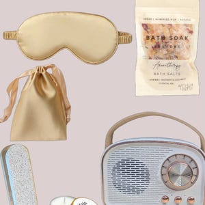 May include: A self-care gift set featuring a white retro radio, a gold satin sleep mask, a small gold satin bag, a wooden foot file, a small candle, a wooden brush, and a bag of bath salts with the text "Bath Soak Restore" and "Aromatherapy Bath Salts".