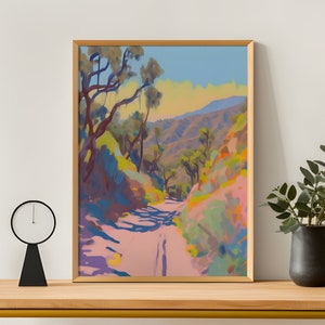 May include: A framed landscape painting shows a road through a canyon, bathed in sunlight. The artwork uses pink, blue, green, and yellow. Trees line the path. The painting is on a wooden table with a black clock and a vase.