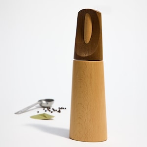 Może przedstawiać: Drewniany młynek do soli i pieprzu z jasnobrązowym korpusem i ciemnobrązową górą. Góra ma unikalny wzór z małym otworem w środku.