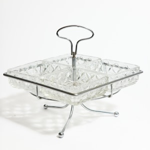May include: A vintage serving dish with four square glass compartments, each with a diamond pattern. The dish is set in a chrome metal frame with a handle and four curved legs. A classic piece for serving snacks.