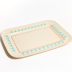 Puede incluir: Bandeja rectangular de color crema con un patrón de triángulos azul claro alrededor del borde y un borde marrón. La bandeja tiene esquinas redondeadas y un diseño sencillo, adecuada para servir alimentos o exhibir objetos.