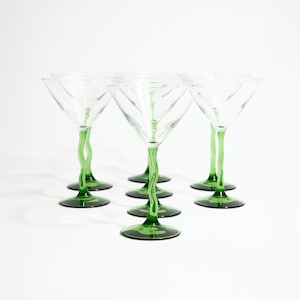 Puede incluir: Juego de seis copas de martini transparentes con tallos y bases verdes. Los tallos tienen un diseño retorcido único. Las copas están dispuestas sobre una superficie blanca, mostrando su elegante forma y contraste de color.