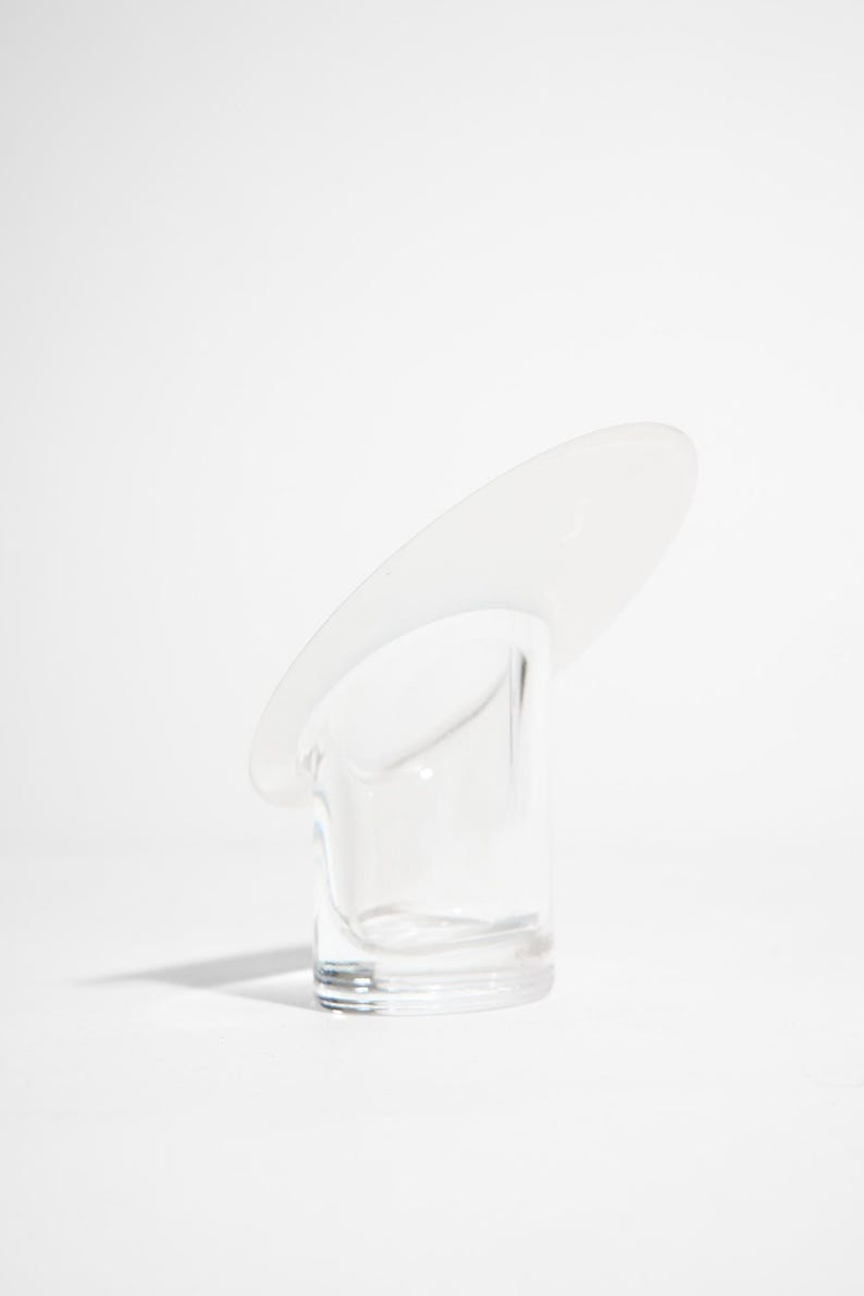 May include: A clear glass object with a cylindrical base and a curved, oval-shaped top. The object is transparent, allowing light to pass through. The design is minimalist and modern, set against a white background.