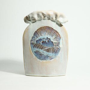 Puede incluir: Un jarrón de cerámica con un esmalte azul claro y blanco. El jarrón tiene una forma única y presenta un diseño pintado a mano de un sol saliendo sobre las olas.