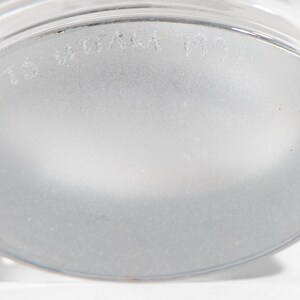 May include: Close-up of the frosted base of a clear glass. The glass has a rounded base and a slightly textured surface. The text "TS WOA" is visible on the base.