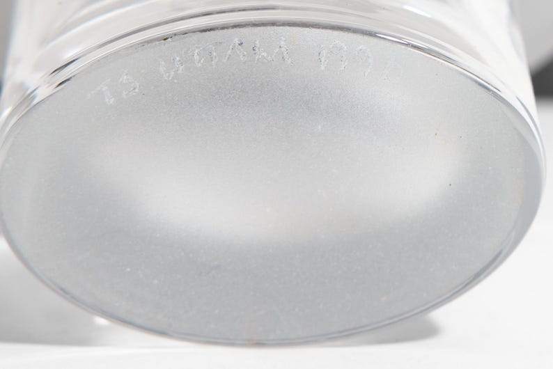 May include: Close-up of the frosted base of a clear glass. The glass has a rounded base and a slightly textured surface. The text "TS WOA" is visible on the base.