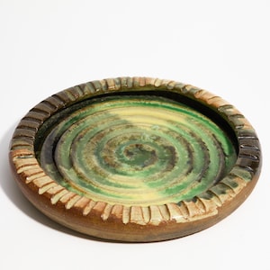 Puede incluir: Un cenicero redondo de cerámica con un esmalte verde y marrón. El cenicero tiene un borde elevado con un diseño texturizado.