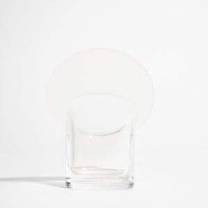 May include: A clear glass votive candle holder with a circular, opaque white disc attached to the top. The candle holder is square-shaped and sits on a white surface. The design is minimalist and modern.