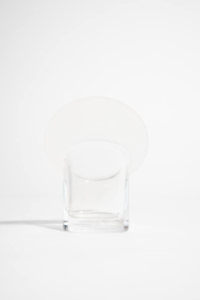 May include: A clear glass votive candle holder with a circular, opaque white disc attached to the top. The candle holder is square-shaped and sits on a white surface. The design is minimalist and modern.