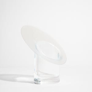 May include: A clear glass vase with a circular, tilted top. The vase has a cylindrical base and a frosted, oval-shaped opening on the top. The design is minimalist and modern, suitable for displaying flowers or as a decorative piece.