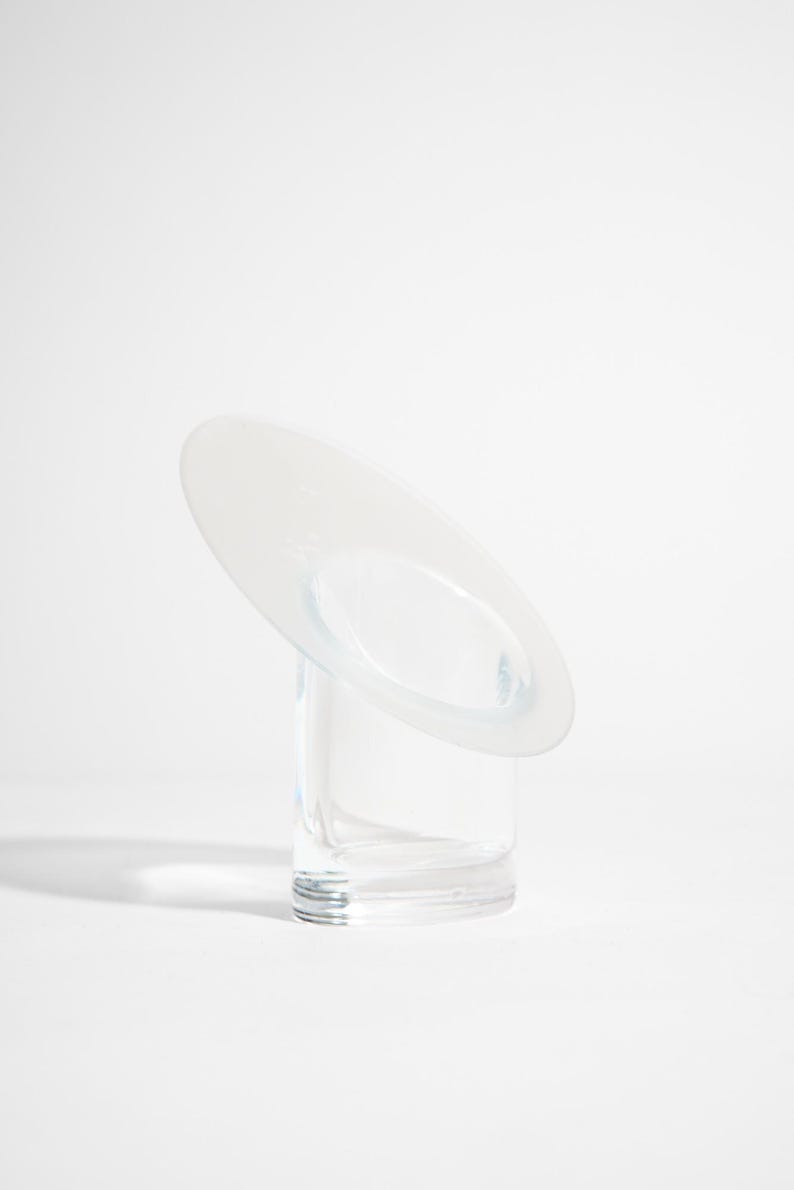 May include: A clear glass vase with a circular, tilted top. The vase has a cylindrical base and a frosted, oval-shaped opening on the top. The design is minimalist and modern, suitable for displaying flowers or as a decorative piece.