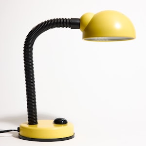 Puede incluir: Una lámpara de escritorio amarilla con cuello y base negros y flexibles. La lámpara tiene una pantalla redondeada y un botón de encendido/apagado negro. La lámpara está sobre un fondo blanco.