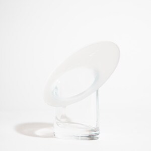 May include: A contemporary glass sculpture with a minimalist design. It features a clear, rectangular base and a white, oval-shaped top with a central opening. The sculpture is set against a white backdrop, emphasising its clean lines.