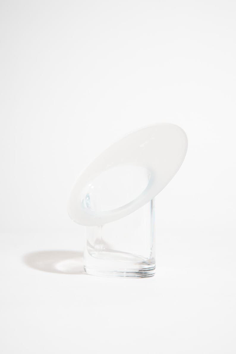 May include: A contemporary glass sculpture with a minimalist design. It features a clear, rectangular base and a white, oval-shaped top with a central opening. The sculpture is set against a white backdrop, emphasising its clean lines.