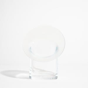 May include: A clear glass vase with a circular top and a rectangular base. The vase has a minimalist design and is set against a white background. The circular top is wider than the base.