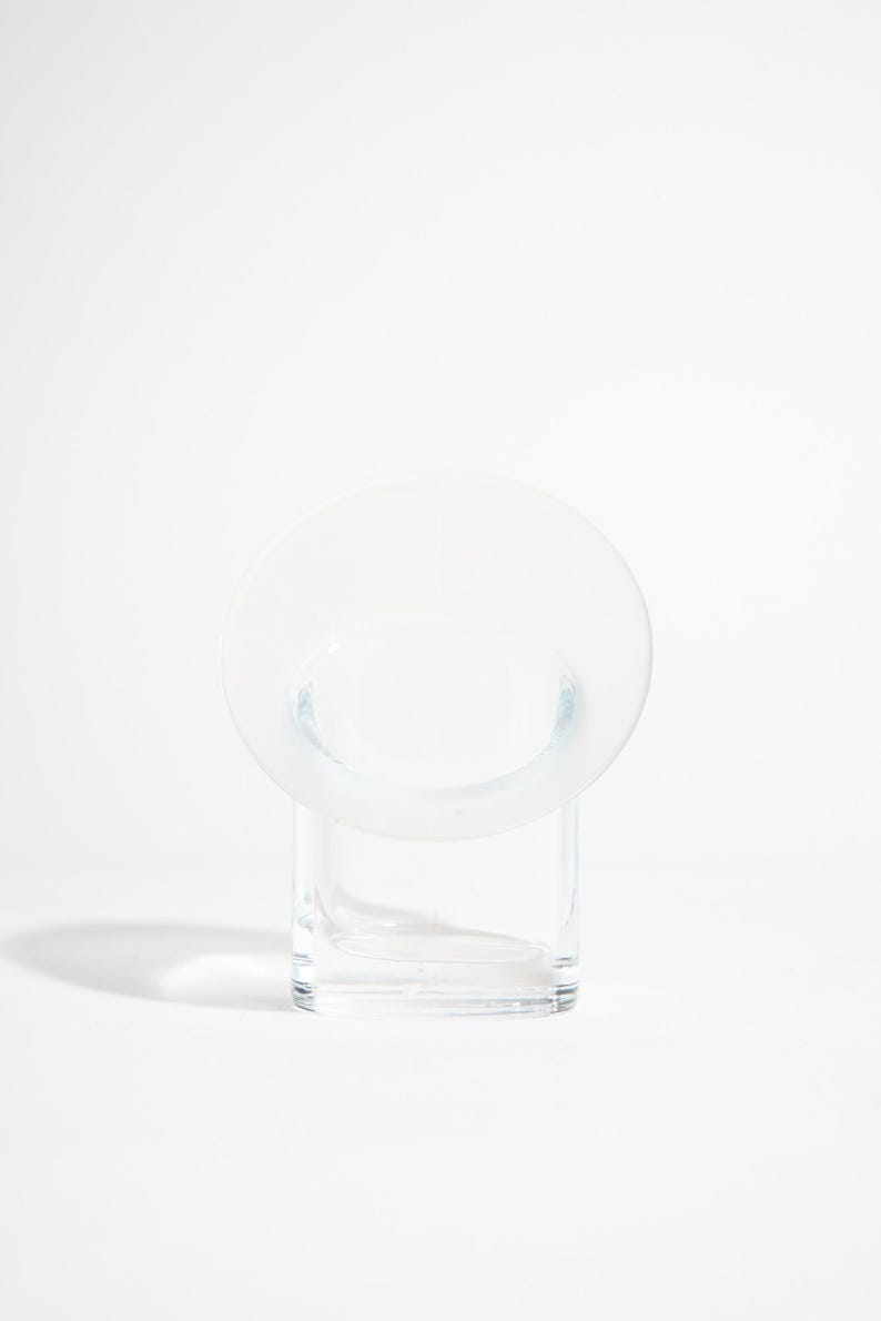 May include: A clear glass vase with a circular top and a rectangular base. The vase has a minimalist design and is set against a white background. The circular top is wider than the base.