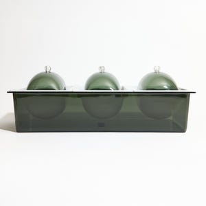 May include: A dark green glass serving dish with three individual compartments, each covered by a matching dome-shaped lid with a small silver handle. The dish is rectangular with a black rim and sits on a white surface.