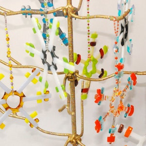 May include: A gold metal stand with multiple branches holds a collection of colorful glass snowflake ornaments. The snowflakes are made of various colors, including red, orange, yellow, green, blue, and white. Each snowflake is unique in its design and shape.